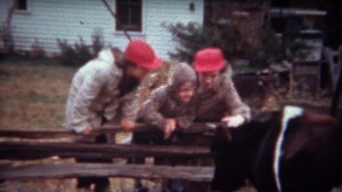 1946: Family in red hats petting cow at rural farmstead.
