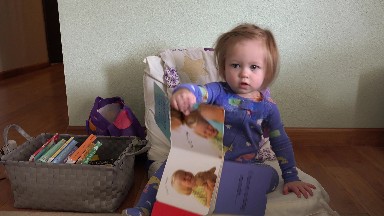 Baby holding up book to read sitting on tiny sized chair in pajamas. DENVER,