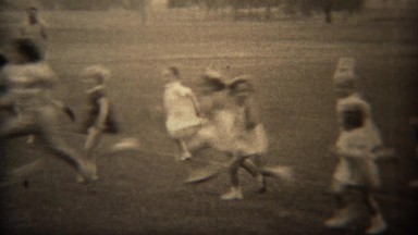 1937: Young girls foot race across grass park field in formal white dress.