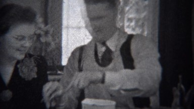 1944: Man wearing apron serving mother food at formal dining table. MIAMI,