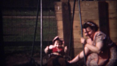 1948: Mother helping pink hood girl child on playground swingset.