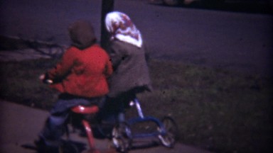 1948: Kids riding tricycles classic cars on neighborhood sidewalk.