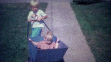 1962: Boy pushing baby in yard waste cart on suburban sidewalk.
