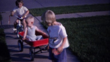 1962: Kids pull red wagon ride tricycle outdoor summer play.