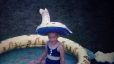 1963: Girl wears inflatable water raft on head in kiddie pool.