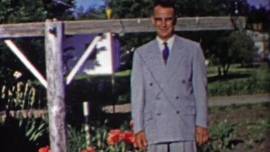 1958: Dapper man in gray double breasted suit posing by flowers.
