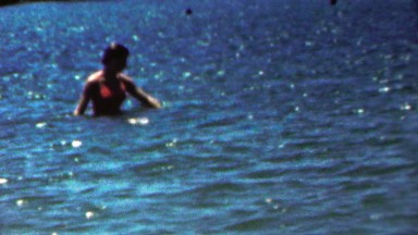 1961: Bikini clad woman wading in deep glistening blue ocean water.