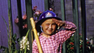 1964: Grade school aged kid salutes military march fireman hat costume.