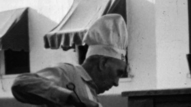 1964: Big chef hat middle aged man cooking outdoor fire working.