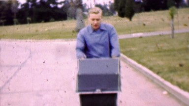 1957: Dad practicing pushing baby stroller in tropical cemetery park.