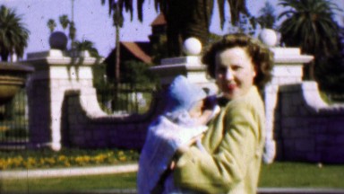 1957: Enthusiastic mother bouncing newborn baby outdoor palm trees.