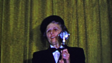 1957: Woman solo sarcastic comedy routine behind old microphone.