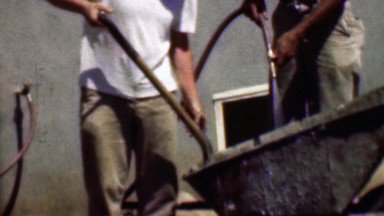 1957: Construction workers mix concrete in wheelbarrow with toddler.