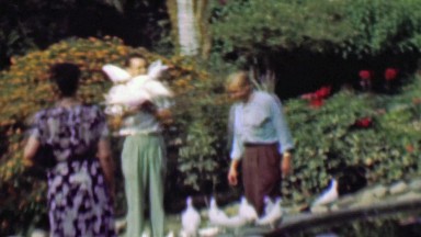 1958: Lunatic man covered in white pigeon birds gets passed by.