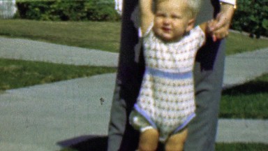 1958: Dad helps baby boy learn walk front yard suburban sidewalk.