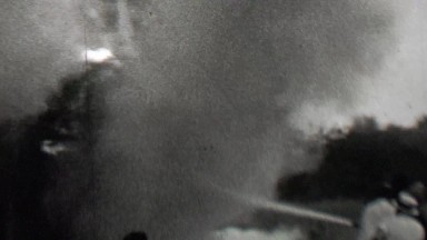 1939: Burning building firemen water hose spray smoke crowd watches.