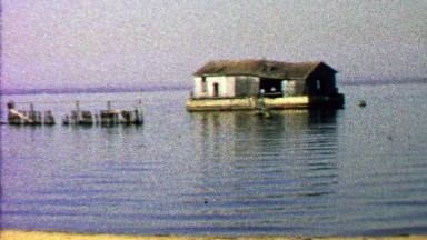 1967: Abandoned ramshackle house floating calm lake waters.