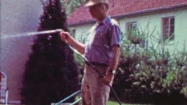 1965: Man sprays camera lense with garden water hose.