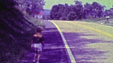 1968: Boy walking to 1st day of school down country road waves goodbye.