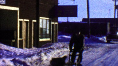 1957: Sled dog pulls man small town winter cold weather.