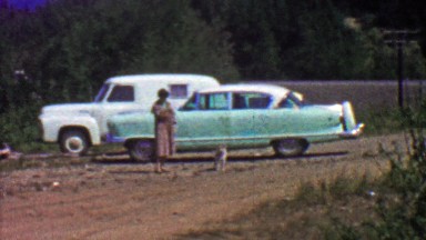 1957: Woman holding leashed dog outside classic cars.