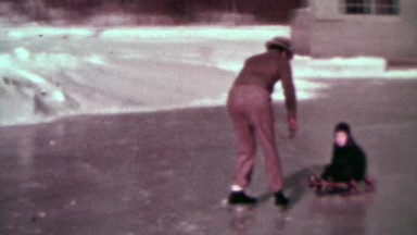 1937: Man ice skating pulling kid on winter snow sled around homemade rink.