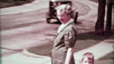1937: Grandma walking suburban toddler boy as classic cars drive by.