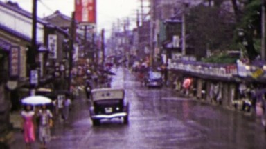 1951: Busy Japanese commercial street raining umbrellas drawn.