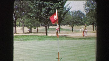 1968: Left handed man hitting golf ball from sand trap onto green near pin hole.