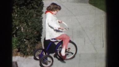 1958: Girl riding tricycle bike on residential sidewalk on sunny winter day.