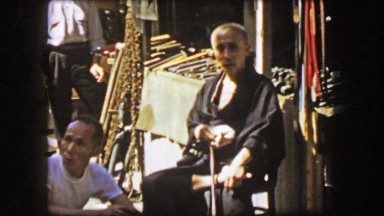 1944: Wise Asian man shining violin instrument on street corner market.