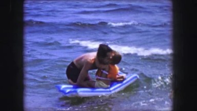 1955: Dad holds happy toddler son inflatable raft ocean wave waters.