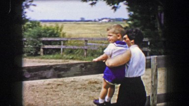 1956: Mother toddler son watching horse ranch riders in stable pens.