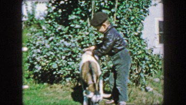 1957: Boy petting tied up farm goat on green grass acred country villa.