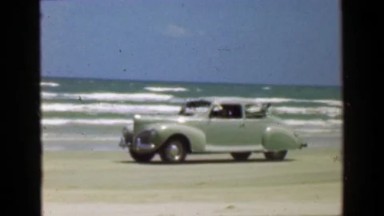 1952: Car driving on ocean beach sand in front of crashing surf waves. PORT
