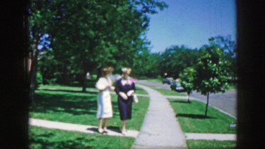 1961: the scene shows 2 women holding together and walking beside a street