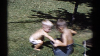 1959: young boy sitting in a tin pail splashes cup filled with water on toddler