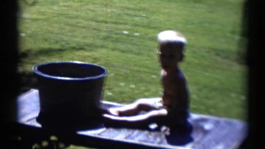 1959: young boy pulling baby on a wagon without sides with a bucket on the wagon