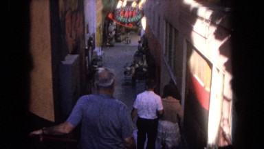 1962: two men and a lady walking down a narrow alley towards a cafe sign