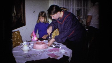 1973: a woman cutting a pink birthday cake as a child attempts to put on a party