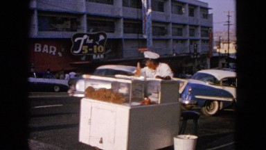 1963: street food seller watching cars passing by while two white men cross the