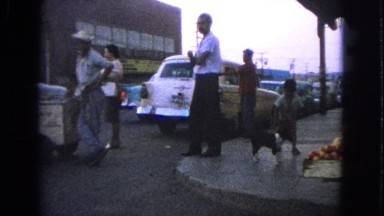 1963: a group of people and a dog mingling on the sidewalk with traffic