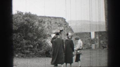 1952: a woman a long coat approaches a threesome standing on a lawn in front of