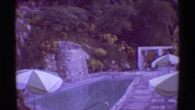 1971: The scenery of a forest with a view of a swimming pool, umbrellas, houses,