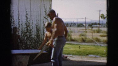 1968: an adult man teaching two children to use a hand saw while cutting a board