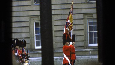 1969: uniform men lining up in formation as onlookers watch from beyond a fence