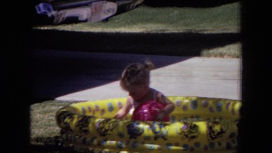 1970: a little girl playing with a red ball as she sits in a yellow kiddie pool