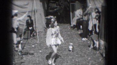 1948: female performer dressed in short skirt walks through outdoor encampment