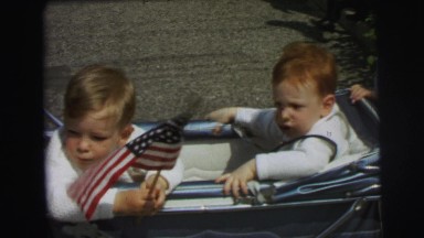 1972: Two Cute Kids With U.S. Flag Sitting in a Trolley Beside Their Mom