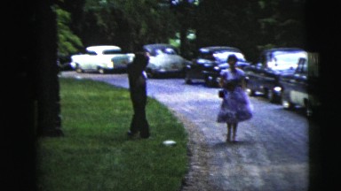 1962: man and woman dressed in formalwear walk next to a row of parked cars,
